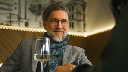 Mature elegant man sits in modern cafe with woman, drinks wine and talks. Couple on date in restaurant. Husband and wife celebrate marriage anniversary, spend leisure time. Concept of public catering.