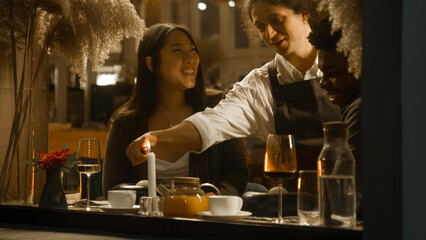 African american man and asian woman sit in modern gastro cafe together, drink wine and talk. Male waiter puts dishes on table. Multi ethnic couple on a date in restaurant. Concept of public catering.
