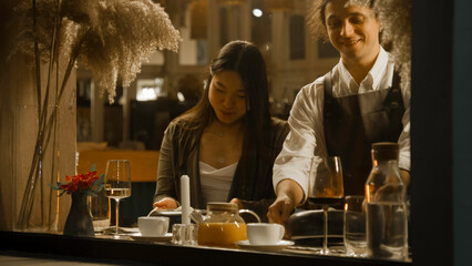 African american man and asian woman sit in modern gastro cafe together, drink wine and talk. Male waiter puts dishes on table. Multi ethnic couple on a date in restaurant. Concept of public catering.