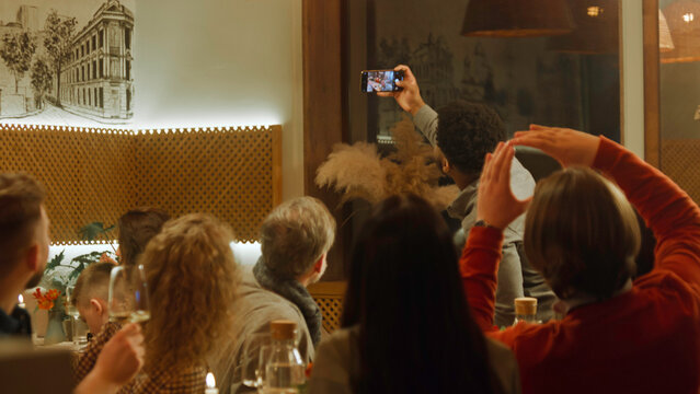 African American man takes photo and selfie with friends in cozy cafe using phone. Group of diverse people spend evening together, hangout on birthday party in restaurant. Concept of public eating. - Powered by Adobe