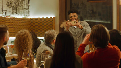 African American man takes photo and selfie with friends in cozy cafe using phone. Group of diverse people spend evening together, hangout on birthday party in restaurant. Concept of public eating.