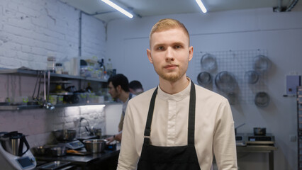 Professional chef in apron stands by workplace and looks at camera. Busy coworkers on background preparing dishes. Concept of teamworking and cooking at restaurant kitchen. Portrait view. Slow motion.