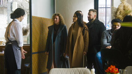 Male waiter greets friends company on entrance in restaurant with modern design and sits them at the reserved table. Multi ethnic couple on a romantic date at background. Concept of public eating.