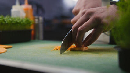 The cook cuts sweet potato on cutting board. Hands of professional chef close-up. Greenery and other ingredients on kitchen desk. Process of cooking gourmet dish. Public catering. Vegetarian dish.