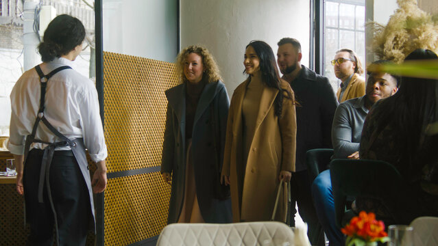 Male waiter greets friends company on entrance in restaurant with modern design and sits them at the reserved table. Multi ethnic couple on a romantic date at background. Concept of public eating.