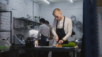 Male cook in apron stands by kitchen table and cuts sweet potato. Coworker on background peels vegetables for dishes. Professional chef preparing food. Concept of working in restaurant. Slow motion.