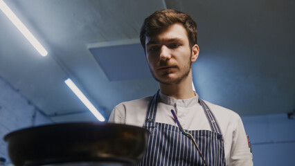 Male cook in apron fries and stirs juicy meat in frying pan over stove. Process of preparing...