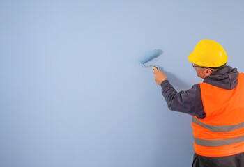 A painter in overalls and a helmet paints a light blue wall with a paint roller. copy space muck-up