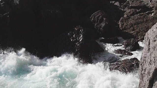 Slow motion. View from above. Brightly turquoise waves break into white foam on the coastal cliffs and seethe between them, sparkling in the rays of the summer sun.