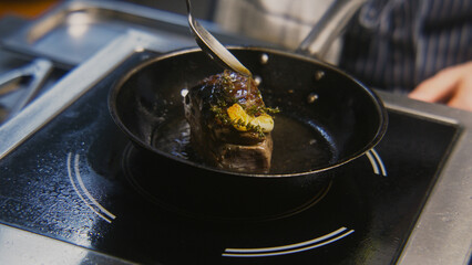 Chef cooks tasty meal on stove. Male cook fries juicy meat steak on pan with rosemary and garlic, pours beef meat with oil. Process of roasted steak cooking. Restaurant with professional cuisine.