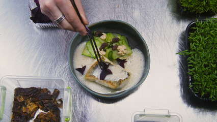 Male cook makes presentation of dish. The chef decorates dish of fried fish and stewed vegetables in bechamel sauce with flower petals on top. Restaurant with professional gourmet cuisine. Close up.