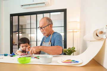 Asian Japanese Senior man and little child boy in apron painting art at home together. Happy Grandfather and Grandson with watercolor.