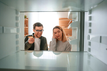 Man is smiling. Woman is complaining about an empty fridge to her husband on the kitchen