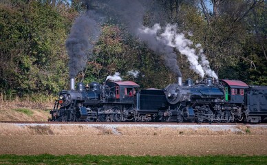 Fototapeta na wymiar View of Two Steam Engines, blowing Smoke and Steam Warming Up Next to Each Other on a Sunny Day