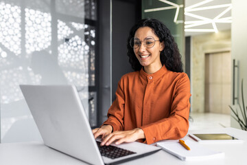 Fototapeta na wymiar Happy and smiling hispanic businesswoman typing on laptop, office worker with curly hair and glasses happy with achievement results, at work inside office building