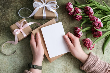 Close-up of female hands holding an envelope with an empty paper postcard on the background of a bouquet of tulips and gifts. The concept of women's day, mother's day, birthday