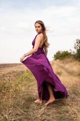 Vertical shot of a Caucasian blonde woman wearing a purple dress posing in a wild nature