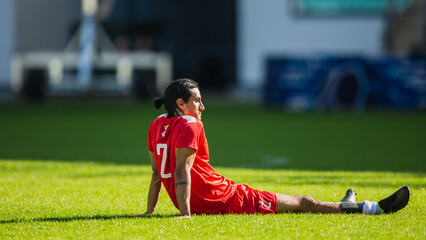 Fototapeta premium International Soccer Football Championship Match: Red Team Player is Sitting on Grass Field of Stadium, Resting During Timeout, Restoring Energy. Footballer Stretching on The Ground.