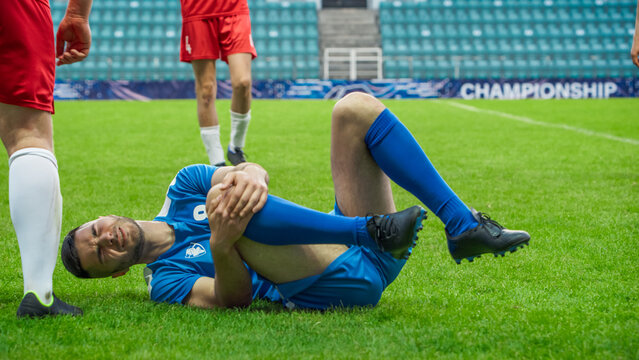 Professional Soccer Football Match Championship: Blue Team Players Attacks, Loses Ball to Foul. Game on an International Tournament. Athlete In Pain Lying on the Grass, Holding a Knee after Falling