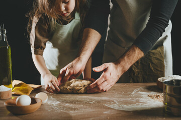 Unrecognizable father and daughter wearing aprons kneading dough on wooden background against dark...