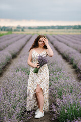Fototapeta na wymiar Beautiful girl in a dress in purple lavender field. Woman walking on the lavender field. Female collects lavender. Enjoy the floral glade, and summer nature. Natural cosmetics and eco makeup concept.