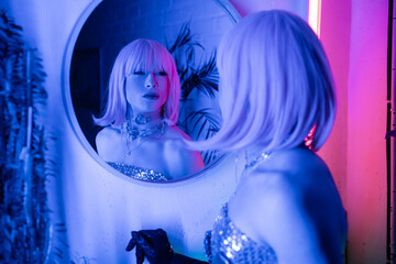 Blurred drag queen in wig and top looking at mirror in neon light at home.