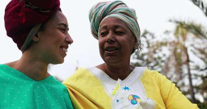 Happy african mother and daughter wearing traditional clothes sitting on meadow with city park in background - Mom day and family concept