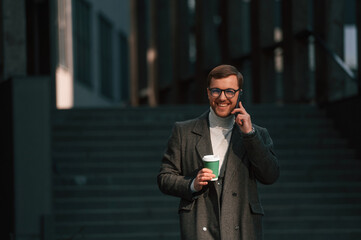 Amazing sunlight, holding smartphone and cup of drink. Handsome man in formal clothes is outdoor near the business building