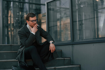 Sitting on the stairs and smoking. Handsome man in formal clothes is outdoor near the business building