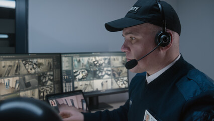 Security operator controls CCTV cameras in office, uses digital tablet and computers, talks using walkie-talkie. Program showing surveillance cameras footage on screens. Monitoring and social safety.