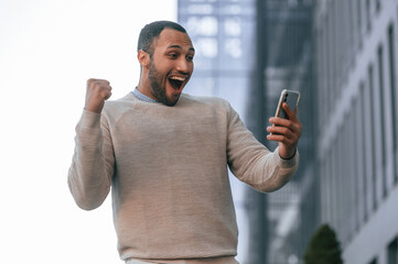 Happy guy holding smartphone and celebrating success. Handsome black man is outdoors near the business building