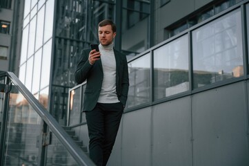 Using social media by smartphone. Handsome man in formal clothes is outdoor near the business building