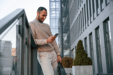 Enjoying social media by using smartphone. Handsome black man is outdoors near the business building