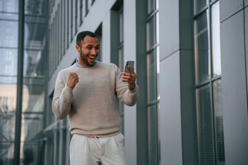 Happy guy holding smartphone and celebrating success. Handsome black man is outdoors near the business building
