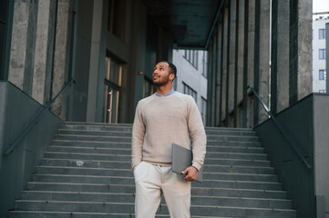 Holding laptop in hand. Handsome black man is outdoors near the business building