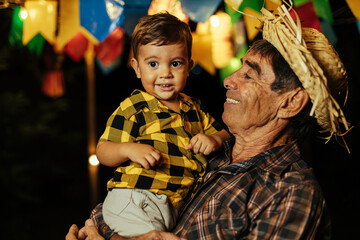 Fototapeta na wymiar Senior man and his baby grandson celebrating the Brazilian Festa Junina. Portrait of grandfather and his grandson wearing typical clothes and a straw hat during the traditional June festival in Brazil