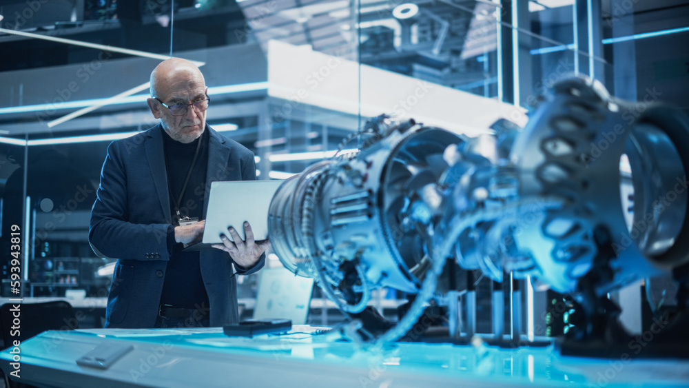 Poster middle aged industrial engineer working on a futuristic jet engine, standing with laptop computer in