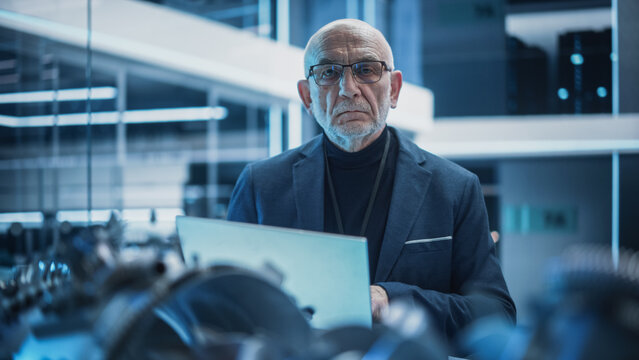 Portrait of a Senior Chief Engineer Using Laptop Computer, Analyzing and Researching a Futuristic Jet Engine. Successful Industrial Scientist in Glasses Looking at Camera.