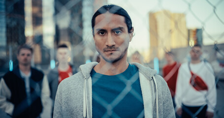 Portrait of a Handsome Multiethnic Man Looking at Camera while Standing Behind a Fence in an Urban City Location. Stylish Diverse Group of Friends Wearing Streetwear Standing Behind Him