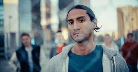 Close Up Portrait of a Focused Multiethnic Man Looking at Camera while Standing Behind a Fence in an Urban City Environment. Stylish Diverse Group of Friends Standing Behind Him