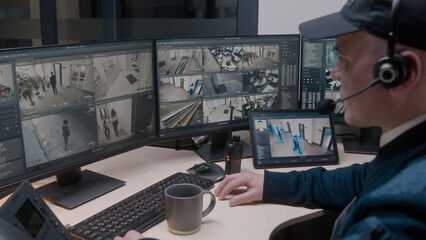 Security officer in headset controls CCTV cameras in office, uses digital tablet and computers....