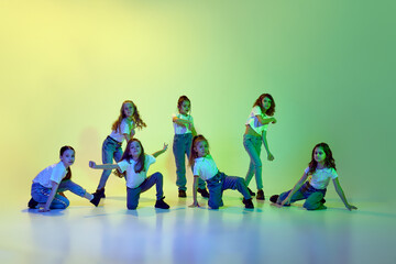 Group of little girls, children in casual clothes dancing, learning modern dance styles against green studio background in neon light. Childhood, hobby, sportive lifestyle, action and motion concept - obrazy, fototapety, plakaty