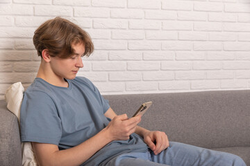 Fototapeta premium Enjoying Weekend. Teenage boy using smart phone sitting on sofa at home