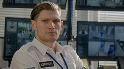 Security worker in uniform sits in observation room and looks at camera. Footage of CCTV cameras on computer multiple monitors at background. Modern program with AI facial scanning system. Portrait.