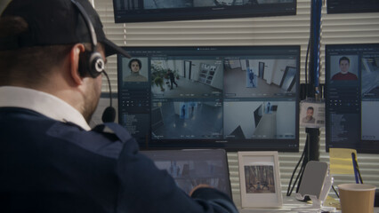 Security officer in headset monitors CCTV cameras on computer in observation room. Multiple PC screens showing footage of surveillance cameras with AI facial recognition high tech software. Back view.