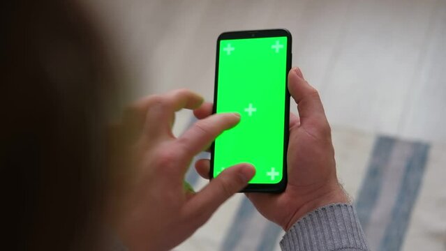 Hipster man sitting at home holding smartphone green mock-up screen in hand. Male person using chroma key mobile phone. Vertical mode. Touching, swiping display, tapping, surfing Internet social media