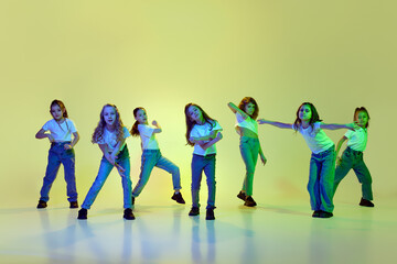 Talented, active, little girls, children dancing hip-hop in sportive casual clothes against green studio background in neon light. Concept of childhood, hobby, sportive lifestyle, active and motion