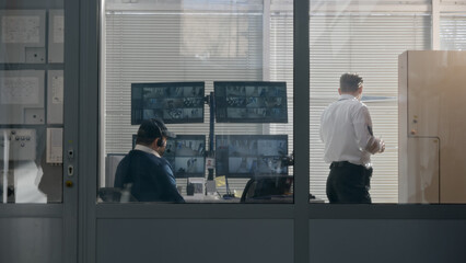 Security worker watches out window through blinds in observation room. Then monitors CCTV cameras displayed on computer screens together with coworker. Observation and tracking system. Social safety.
