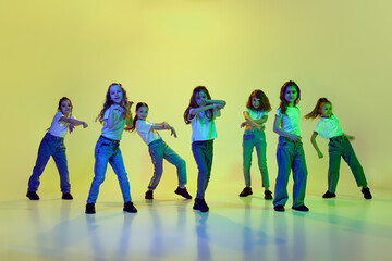 Dynamic image of little girls, children dancing against green studio background in neon light. Casual clothes. Hip-hop style. Concept of childhood, hobby, sportive lifestyle, action and motion