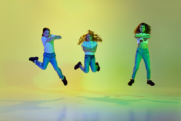 Fototapeta na wymiar Three active, little girls, children jumping, dancing in casual clothes against green studio background in neon light. Concept of childhood, hobby, sportive lifestyle, fashion, action and motion